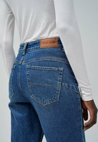 Tamne traperice tamnoplave s kožnom oznakom "Salsa Jeans" na pojasu. Karakteristike uključuju dva stražnja džepa i kontrastne šavove.