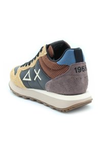 Sneaker sportivo con una tomaia in suede beige e pelle scura, accenti rigati marroni, linguetta blu sul tallone e suola in gomma. Presenta un dettaglio con logo sul lato.