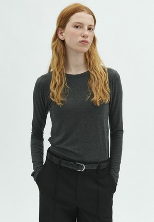 Jong persoon met lang rood haar, gekleed in een donkergrijs shirt met lange mouwen en een zwarte broek, handen in de zakken, tegen een effen achtergrond.
