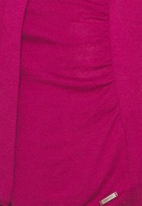 Fuchsia gestricktes Oberteil mit geraffter Textur auf einer Seite. Verfügt über ein kleines silbernes Logo-Täfelchen auf der rechten unteren Seite. Glatte Stoffoberfläche.
