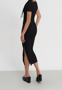 Kvinna i en figurnära svart midiklänning med en sidoklyvning och svarta plattformsmuleklackar, stående mot en enfärgad ljus bakgrund.