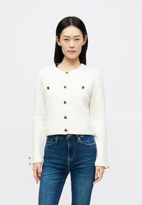 Suéter cropped de mistura de lã branca com decote redondo, botões dourados e dois bolsos no peito. Combinado com jeans skinny azul-escuro.