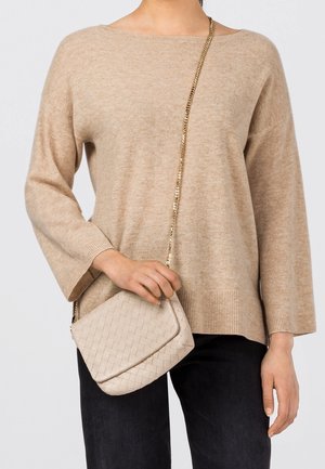 Personne portant un pull beige et un pantalon noir tient un sac à bandoulière en cuir tressé beige avec une bandoulière en chaîne dorée.
