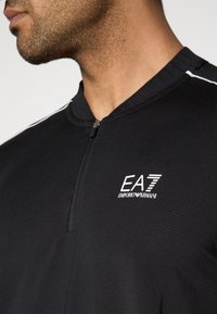 Μαύρο αθλητικό πουκάμισο με φερμουάρ, κατασκευασμένο από υφασμάτινη υφή, που διαθέτει λευκό λογότυπο EA7 στο στήθος και αντίθετες λευκές λεπτομέρειες στους ώμους.