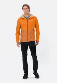 Buitenjas in oranje met een lichter gekleurde capuchon, ritsluiting en zakken. Gecombineerd met zwarte broeken en sportieve schoenen.