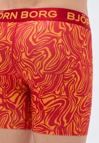 Κόκκινα και πορτοκαλί μποξεράκια με κυματιστό μοτίβο, που διαθέτουν κόκκινη μέση με την επιγραφή "BJÖRN BORG" σε κίτρινα γράμματα. Λείο ύφασμα.