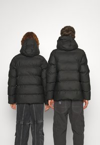 Crni puffer jakne s kapuljačama, kvadratnim dizajnom i širokim rukavima. Nose ih dvije osobe, s vidljivim bočnim ventilacijskim džepovima.