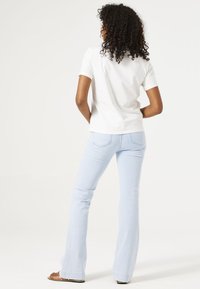 Weißes Kurzarmhemd kombiniert mit hellblauen Schlagjeans. Die Jeans haben Gesäßtaschen und eine dezente Textur. Sandalen mit flacher Sohle.