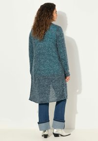 Lång, teal-f�ärgad stickad kofta med texturerad yta. Öppen rygg med sidoklyftor, parat med blå denim och silverfärgade ankelstövlar.