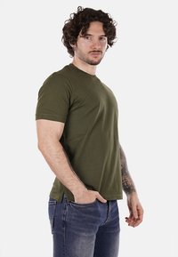 T-shirt verde oliva a maniche corte con scollatura a girocollo, realizzato in tessuto morbido, abbinato a jeans blu, indossato da un uomo con capelli ricci.