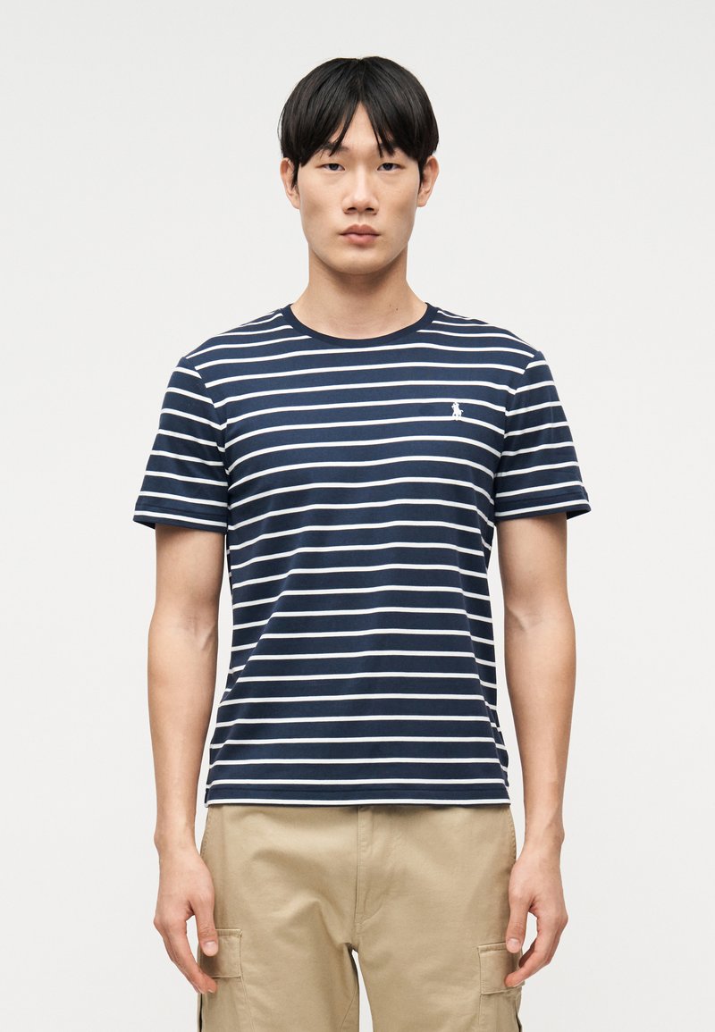 Νεαρός άνδρας που στέκεται αντιμετωπίζοντας προς τα εμπρός, φορώντας μπλε ναυτικό και λευκό ριγέ T-shirt και μπεζ cargo σορτς, μπροστά σε απλό φόντο.