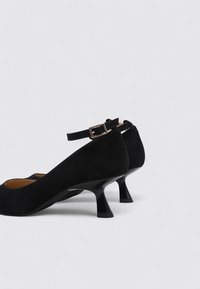 Chaussures en suede noir avec un bout arrondi et une sangle au niveau de la cheville, présentant un petit talon évasé et un intérieur lisse. Design minimaliste.