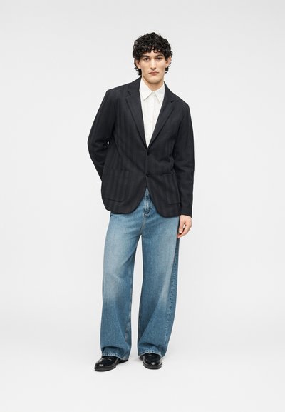 Blazer sombre à motif avec deux poches, porté sur une chemise blanche à col. Jeans denim bleu clair, amples, et chaussures noires complètent le look.