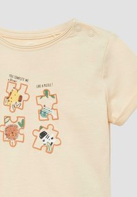 Beżowa koszulka dziecięca z czterema kawałkami puzzli przedstawiającymi żyrafę, kota, lwa i zebry oraz napisem "Uzupełniasz mnie jak puzzle!"