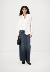 Blusa blanca texturada con mangas globo, combinada con una falda maxi de denim de cintura alta que presenta una abertura frontal. Bolso negro y zapatos.