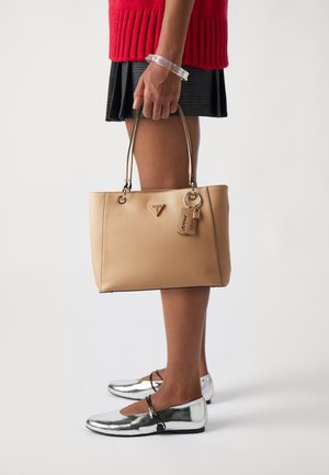 Sac cabas beige avec deux poignées, texture lisse, détail logo et un charm. Le mannequin porte des chaussures plates métalliques argentées et une jupe texturée.