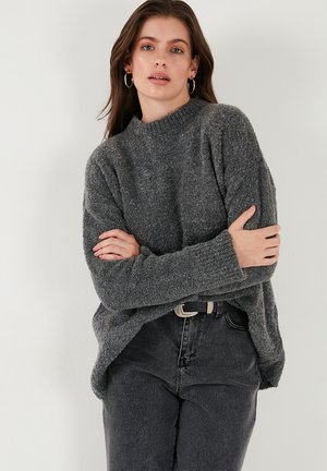 Maglione grigio con texture, collo alto e polsini a coste, abbinato a jeans neri a vita alta e una cintura con fibbia rettangolare.