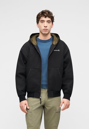 Jeune homme portant une veste à capuche Timberland noire sur un pull bleu et un pantalon cargo kaki, debout devant un fond uni.