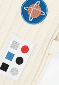 Beige-tilkkutyö, jossa on sininen pyöreä paikka, jossa on oranssi planeetta, sekä valkoinen paikka, jossa on värikkäitä neliöitä ja ympyröitä punaisena, mustana ja sinisenä.