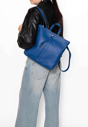 Femme portant une veste en cuir noire et un jean clair, avec un grand sac à dos en cuir bleu porté sur une épaule, sur fond blanc.