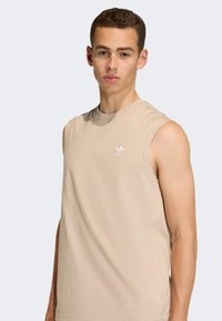 Bež majica bez rukava od mekanog materijala, s okruglim izrezom i malim bijelim logotipom na prsima. Jednostavan i minimalistički dizajn.