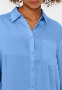 Camicia button-up azzurro chiaro realizzata in tessuto morbido con colletto, taschino sul petto e trama sottile, con bottoni abbinati.