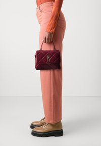 Sac à main matelassé rouge avec une poignée supérieure, arborant un logo en métal doré, tenu à côté d'un pantalon rose clair et de chaussures à lacets beiges.