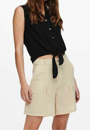Chemise noire sans manches boutonnée nouée à la taille, associée à un short plissé beige taille haute, portée par une personne debout.