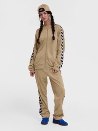 Ženska z vpletenimi lasmi, oblečena v bež trenirko z Chevron vzorcem, modrimi supergami in bež kapo, obrnjeno nazaj, stoji pred belim ozadjem.