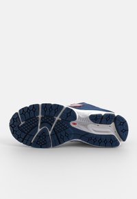 Semelle de basket bleue et blanche avec une bande de roulement texturée et un petit logo rouge sur une surface blanche et un fond gris.