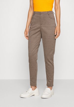 Vrouw draagt bruine geruite taps toelopende broek, witte loafers en een feloranje mouwloze top, staand tegen een effen achtergrond.