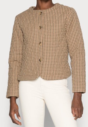 Kvinde iført en beige og brun ternet quiltet jakke med knapper, kombineret med hvide bukser med høj talje, stående mod en ensfarvet baggrund.