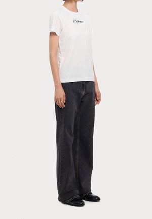 Personne portant un T-shirt blanc avec un texte en écriture noire et un jean noir ample associé à des chaussures noires, debout devant un fond uni.