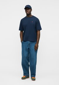 Moški, ki nosi temnomodro kapo in majico, široke modre kavbojke ter večbarvne rjave in zelene čevlje, stoji z rokami v žepih.