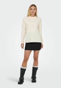 Cremfarbener Rippstrickpullover mit Rundhalsausschnitt, kombiniert mit einem schwarzen Wildleder-Minirock und schwarzen knielangen Stiefeln mit weißen Akzenten und Schnallen.