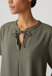 Blusa verde oliva con colletto rotondo e apertura a goccia, caratterizzata da un dettaglio a fiocco al collo e maniche corte. Texture morbida.