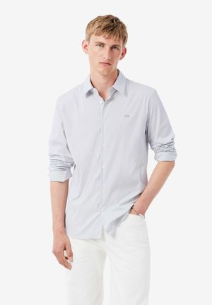 Joven vestido con una camisa de rayas azul claro con las mangas remangadas y pantalones blancos, de pie con una mano en el bolsillo.