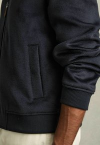 Donker marineblauwe fleece jas met een ritssluiting aan de voorkant, ribgebreide boorden en zoom, en een zijzak op de linkeronderzijde van de voorkant. Zachte textuur.