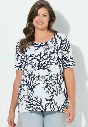 Uśmiechnięta kobieta o falowanych brązowych włosach, ubrana w biały krótki top z wzorem czarnych koralowców i rozgwiazd, w połączeniu z jasnoniebieskimi jeansami.