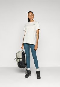 Camiseta de manga corta color beige claro, mallas teal, botas de senderismo negras y una mochila gris con detalles negros. Diseño simple con logotipos.