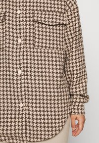 Hnědá a béžová košile s vzorem houndstooth, která má dvě náprsní kapsy a zakřivený lem, vyrobená z texturované tkaniny.