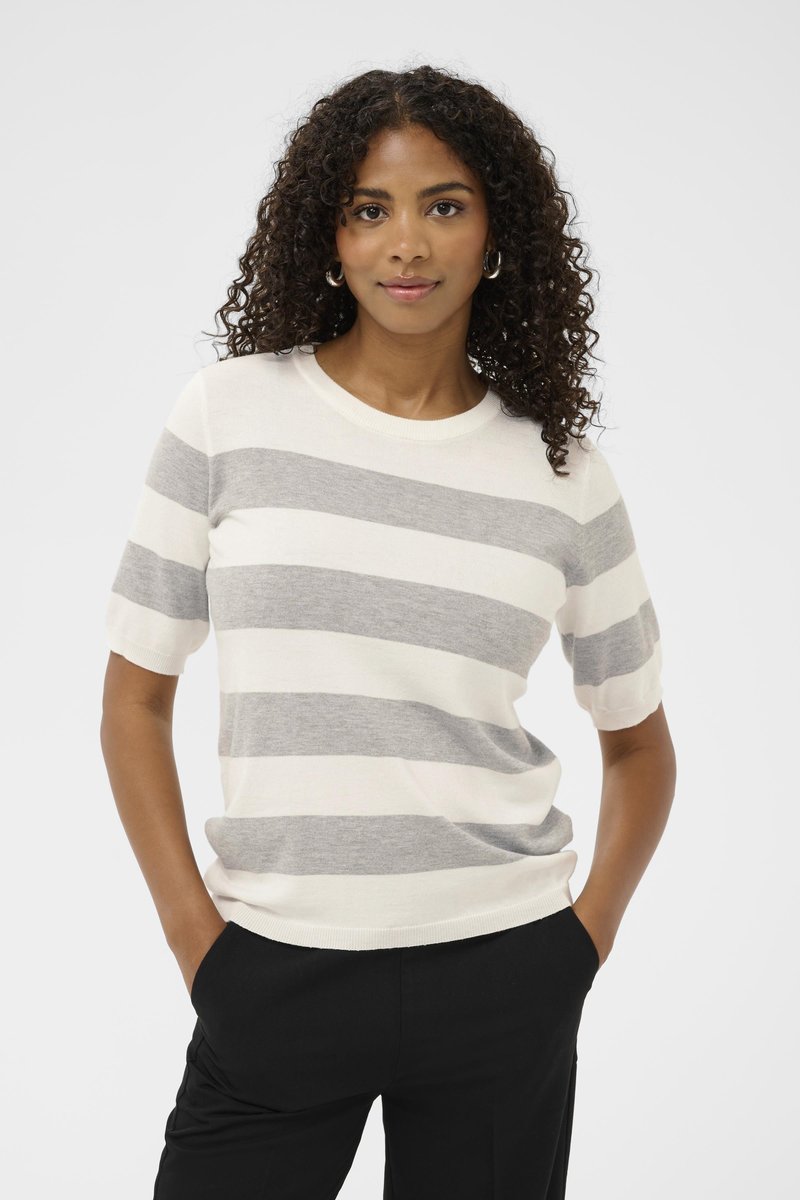 Woman with curly hair wearing silver hoop earrings, a short-sleeve white and gray striped sweater, and black pants, hands in pockets.