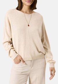 Beige Strickpullover mit langen Ärmeln, lockerer Passform, Rundhalsausschnitt und geripptem Saum. Kombiniert mit hellen Hosen und einer goldenen Halskette.