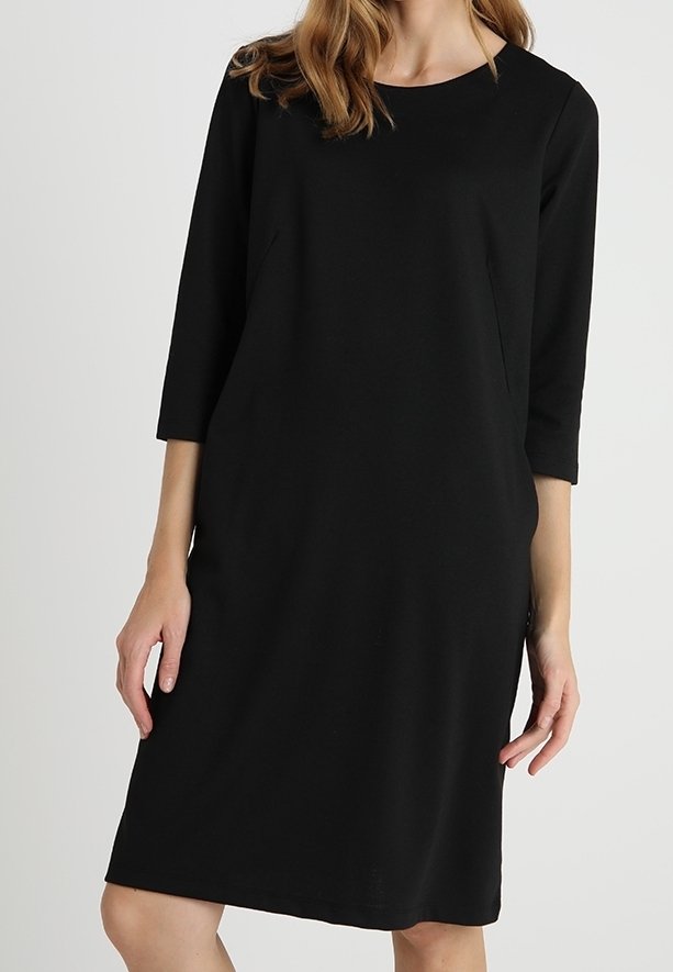 Femme portant une robe noire longueur genoux, manches longues, avec un col rond, se tenant devant un fond clair uni.