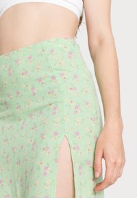 Saia floral verde com pequenas flores cor-de-rosa e amarelas. Apresenta uma fenda lateral e um tecido leve. Close-up na cintura e no braço.