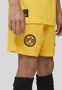 Pantalones cortos de fútbol amarillos con tejido texturizado; presenta un logo negro de BVB 09 y una etiqueta negra de Puma en el costado. Medias negras exhibidas.