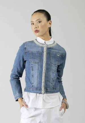 Giovane donna che indossa una giacca di jeans decorata con bordi di perle sopra una camicia bianca con bottoni e pantaloni bianchi, mani nelle tasche, sfondo neutro.