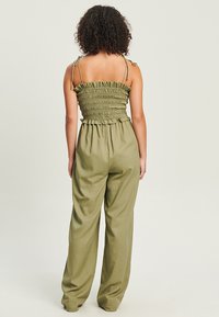 Combinaison en vert olive avec corsage froncé, bretelles réglables, jambes larges et taille froncée, fabriquée dans un tissu léger.