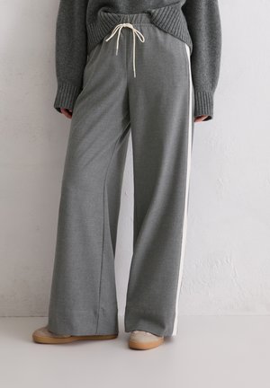 Person, der lockere graue Hosen mit weißen Seitenstreifen, einen grauen Pullover mit langen Ärmeln und beigen Schuhen trägt, vor einer hellen Wand steht.