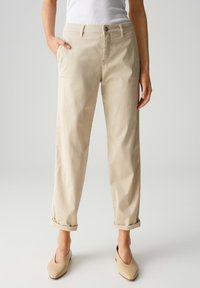 Beige Baumwollhose mit entspannter Passform, ausgestattet mit Gesäßtaschen, umgeschlagenen Säumen und einem Knopfverschluss, kombiniert mit beige-farbenen Slippern.